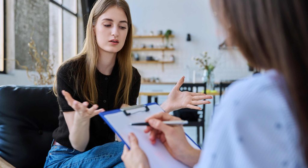 Two people in a casual meeting: a woman speaks and gestures while the other person holds a clipboard.