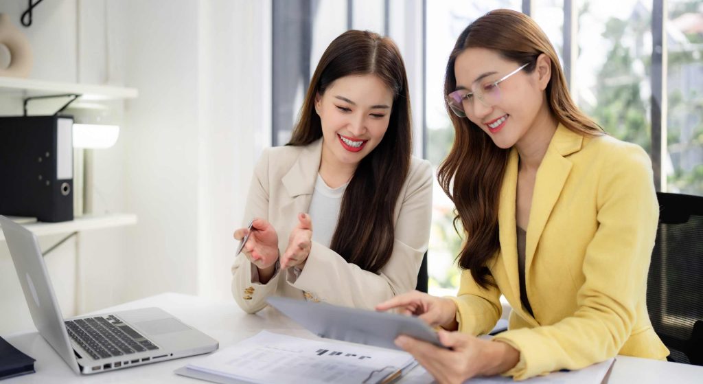 Two women collaborate at a bright desk, smiling while reviewing documents and a laptop nearby.