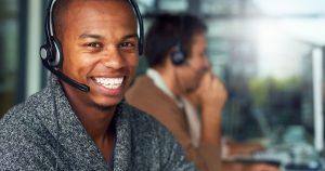 Smiling man wearing a headset in a call center, with another agent in the background.