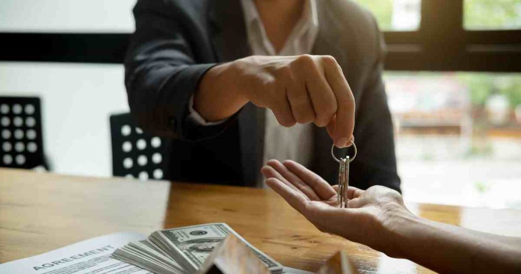 Person in a suit hands over a set of keys to another person across a table, with cash and a signed agreement visible nearby