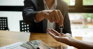Person in a suit hands over a set of keys to another person across a table, with cash and a signed agreement visible nearby