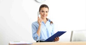Receptionist smiling while talking on a corded phone and holding a blue clipboard at a desk/ reception area