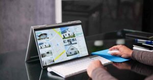 Person using a laptop with a touchscreen displaying a real estate map and property photos on the screen, with a hand on the mouse and keyboard nearby.