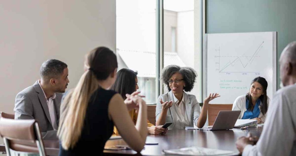 Team of diverse professionals in a meeting around a conference table, one woman speaking with hand gestures while others listen.