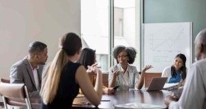 Team of diverse professionals in a meeting around a conference table, one woman speaking with hand gestures while others listen.