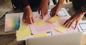 Team collaborating on a paper wireframe with sticky notes and color swatches on a wooden table, planning a design mockup while pointing at layouts.