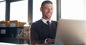 Smiling man in a sweater and tie works on a laptop in a bright office with packages in the background.
