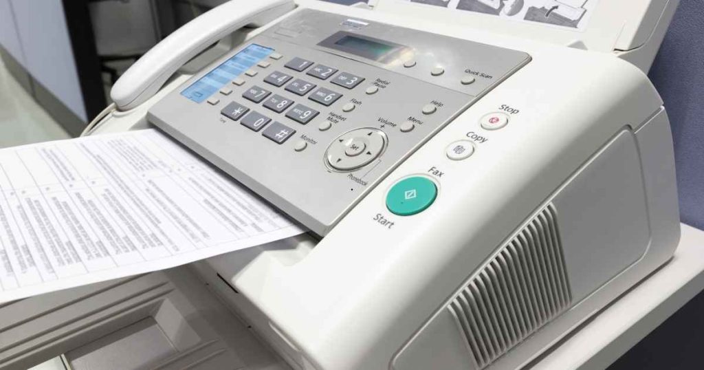 Fax machine with numeric keypad and a printed sheet feeding out of the tray, ready to send or receive a page.